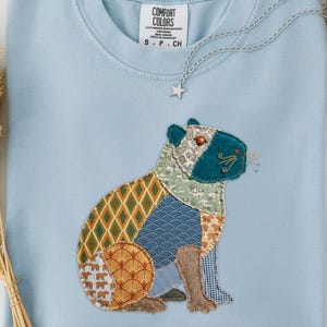 May include: Light blue sweatshirt with a patchwork capybara design. The capybara features various patterned fabrics in shades of green, orange, blue, and brown. The sweatshirt has a crew neck and a small tag that says "Comfort Colors". A silver necklace with a star pendant is draped across the shirt.