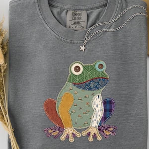 May include: A grey Comfort Colors t-shirt featuring a colorful frog applique. The frog has a patchwork design with various patterns and colors, including green, blue, orange, and purple. A silver chain with a star pendant is draped across the shirt.