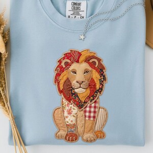 Peut inclure: Un sweat-shirt bleu clair orné d'un appliqué de lion détaillé. Le lion présente un design patchwork avec divers motifs et couleurs, dont du paisley rouge, des imprimés floraux et du vichy. Le sweat-shirt a un col rond.
