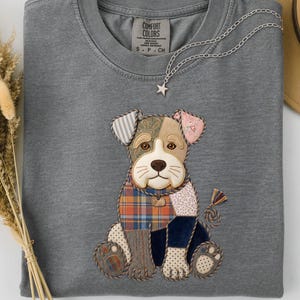 May include: A grey Comfort Colors t-shirt featuring a patchwork dog design. The dog has a brown nose, ears with floral and striped patterns, and a plaid scarf. A silver necklace with a star pendant is draped on the shirt.