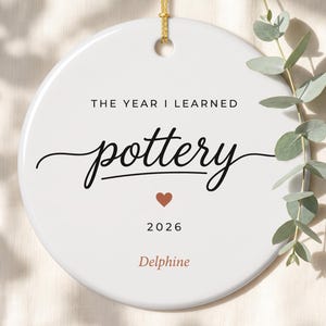 May include: A round white ceramic ornament featuring the black text THE YEAR I LEARNED pottery, a small terracotta heart, the year 2026, and the name Delphine in terracotta script. The ornament hangs from a gold string with a green eucalyptus sprig.