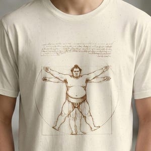 May include: A cream-coloured t-shirt with a sumo wrestler design, inspired by Leonardo da Vinci's Vitruvian Man. The brown illustration shows the wrestler within a circle and square. The t-shirt is worn by a person.