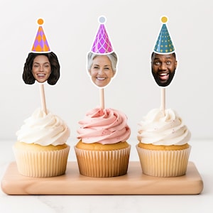 May include: Three cupcakes with white, pink, and white frosting topped with a face cutout wearing a party hat. The hats are orange, purple, and blue with gold stars.
