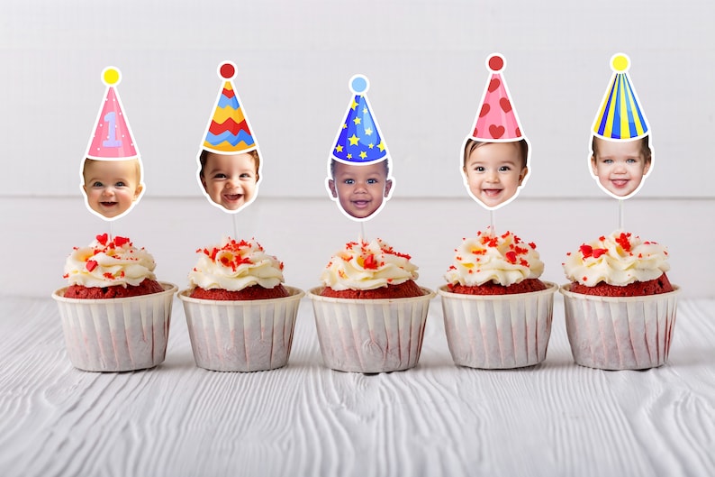 Five cupcakes with white frosting and red sprinkles are topped with baby face cutouts wearing colorful party hats. The hats have various patterns, including a "1" for a first birthday, on a light wooden background.