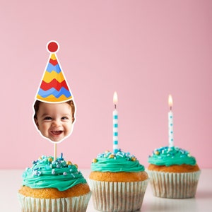 May include: Three cupcakes with teal frosting and colorful sprinkles. One cupcake has a birthday hat with a child's face on it. The cupcakes are on a white surface with a pink background.