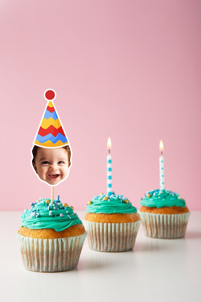 May include: Three cupcakes with teal frosting and colorful sprinkles. One cupcake has a birthday hat with a child's face on it. The cupcakes are on a white surface with a pink background.