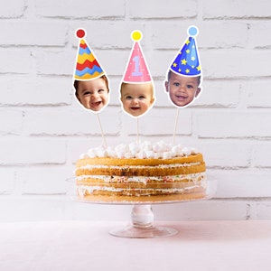 May include: A three-tiered cake decorated with white frosting and topped with three cake toppers featuring photos of babies wearing colorful party hats.