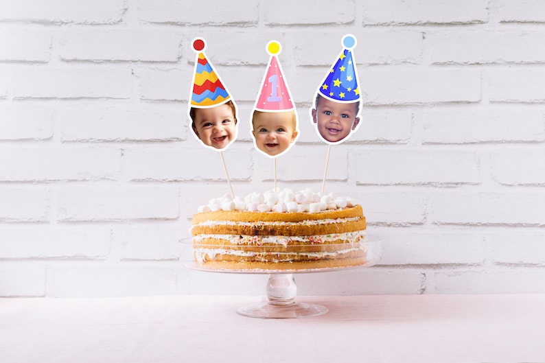 May include: A three-tiered cake decorated with white frosting and topped with three cake toppers featuring photos of babies wearing colorful party hats.