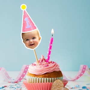 May include: A pink cupcake with pink frosting and sprinkles, topped with a pink and white birthday hat with a baby's face on it and a pink and white candle. The cupcake is sitting on a white surface with pink and blue confetti.