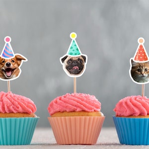 May include: Three cupcakes with pink frosting and colorful animal toppers. The toppers are a dog wearing a blue and pink party hat, a pug wearing a green party hat with white polka dots, and a cat wearing a red party hat with white hearts.