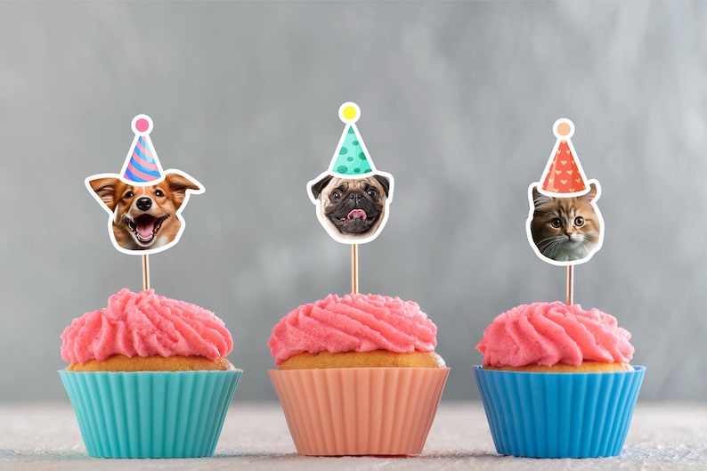 May include: Three cupcakes with pink frosting and colorful animal toppers. The toppers are a dog wearing a blue and pink party hat, a pug wearing a green party hat with white polka dots, and a cat wearing a red party hat with white hearts.