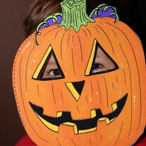 May include: A child wearing a Halloween costume with a pumpkin face mask. The mask is orange with a black smile and eyes. The mask has glitter on it.