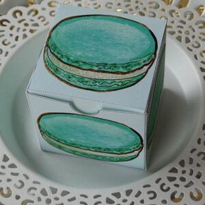 Puede incluir: Una caja de regalo azul claro con una ilustración de acuarela de un macarrón turquesa con relleno blanco y detalles dorados. La caja está colocada sobre un plato blanco decorativo con borde festoneado.