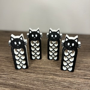 May include: Four black and white 3D printed cat-shaped clips. Each clip has a black cat face with white eyes and a white body with black details. The clips are arranged in a row on a wooden surface.