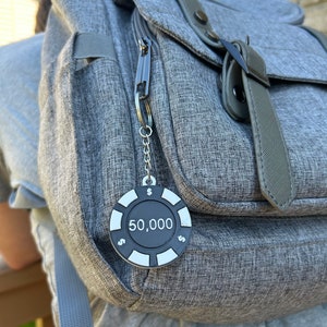 May include: A silver keychain with a black poker chip charm that says "$50,000" hanging from a gray backpack.
