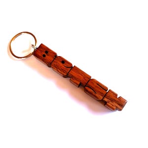May include: A wooden keychain with a segmented figure and a gold-coloured ring. The figure is made of reddish-brown wood, with carved details suggesting a person. The keychain is set against a white background.