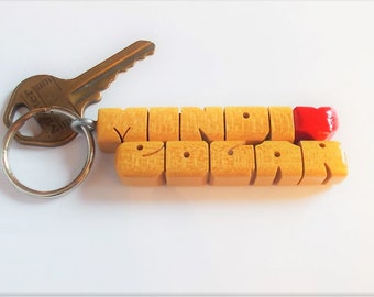 May include: A yellow wooden keychain with the words "MIND YOUR" and a red heart charm. The keychain is attached to a silver key ring and a brass key.