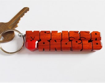 May include: A wooden keychain with the name "Melissa Angelo" carved in a block letter style. The keychain is made of reddish-brown wood and has a red heart-shaped accent. A gold-colored key is attached to the keychain.