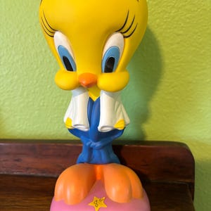 Op de afbeelding: Een gele Tweety Bird-beeldje met blauwe ogen, een oranje snavel en oranje voeten. Het beeldje draagt een blauwe outfit met een witte kraag en staat op een roze voet met een oranje stervormige sticker.