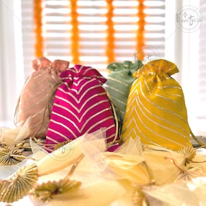 Może przedstawiać: Cztery małe materiałowe torebki prezentowe ze sznurkiem do ściągania. Torebki są w kolorze różowym, zielonym, magenty i żółtym z złotymi paskami. Torebki leżą na białej i złotej powierzchni z tkaniny.