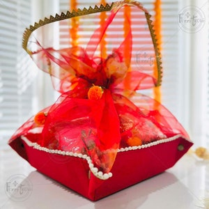 Può includere: Un cesto regalo rosso con base quadrata, adornato con tessuto rosso trasparente e arancione fantasia. Il cesto è rifinito con perle bianche e un manico bordato d'oro. Il cesto è pieno di arance.