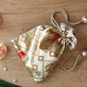 May include: A beige drawstring pouch with a gold geometric pattern and red accents. The pouch is tied with a gold cord and has decorative gold and silver tassels. Pink and yellow flowers and a green leaf are arranged around the pouch on a wooden surface.