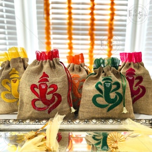 May include: Five small burlap bags with drawstring closures, each decorated with a different colored embroidered image of the Hindu deity Ganesha. The bags are lined with colorful sheer fabric and are arranged on a silver tray.