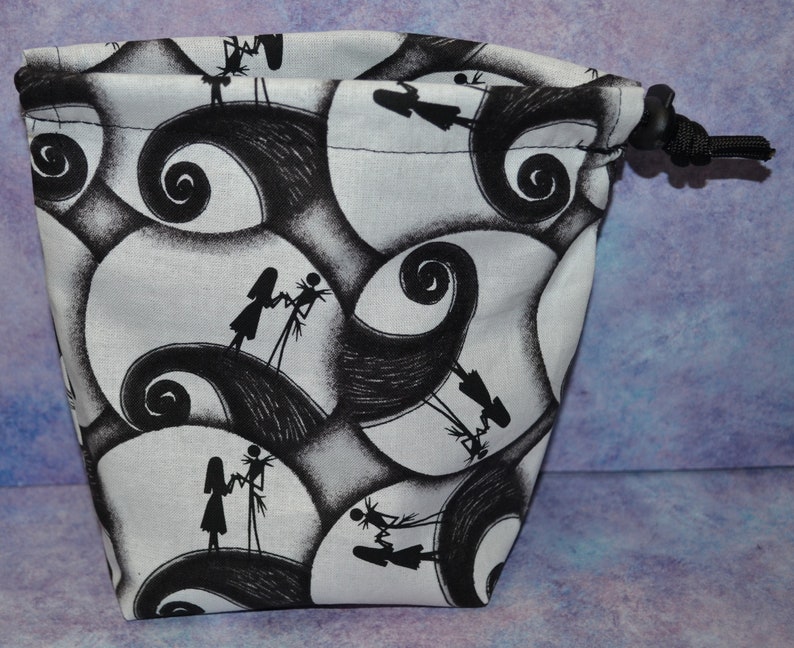 Puede incluir: Una bolsa de cord&oacute;n blanco con un estampado en blanco y negro de Jack Skellington y Sally de Pesadilla Antes de Navidad. El estampado presenta a los personajes caminando sobre un patr&oacute;n de espiral en blanco y negro.