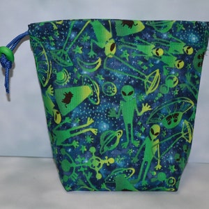 Puede incluir: Una bolsa de cordón verde y azul con un estampado de temática espacial que presenta extraterrestres, naves espaciales y planetas. La bolsa tiene un cierre de botón verde.