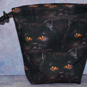 May include: Black drawstring bag with a repeating pattern of black cats with orange eyes. The bag has a black drawstring closure.