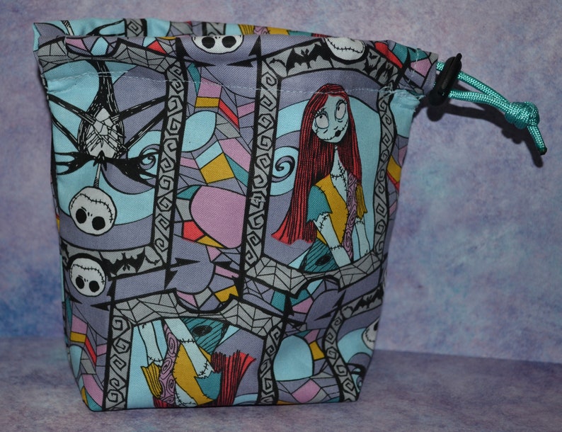 Puede incluir: Una bolsa de cord&oacute;n azul con un estampado de vitral colorido con personajes de Pesadilla antes de Navidad. El estampado incluye a Sally, Jack Skellington y murci&eacute;lagos.