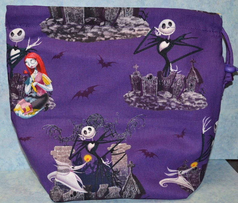 Puede incluir: Bolsa de cord&oacute;n morado con un patr&oacute;n repetido de Jack Skellington y Sally de Pesadilla Antes de Navidad. Los personajes se representan en un cementerio con murci&eacute;lagos volando en el fondo.