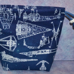 May include: A blue drawstring bag with a white print of various spaceships. The bag has a white drawstring and a white toggle.
