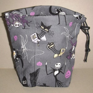 Puede incluir: Una bolsa de cord&oacute;n gris con un estampado en blanco y negro de personajes de Pesadilla antes de Navidad. La bolsa tiene un cord&oacute;n negro y un estampado morado y blanco de calabazas y murci&eacute;lagos.