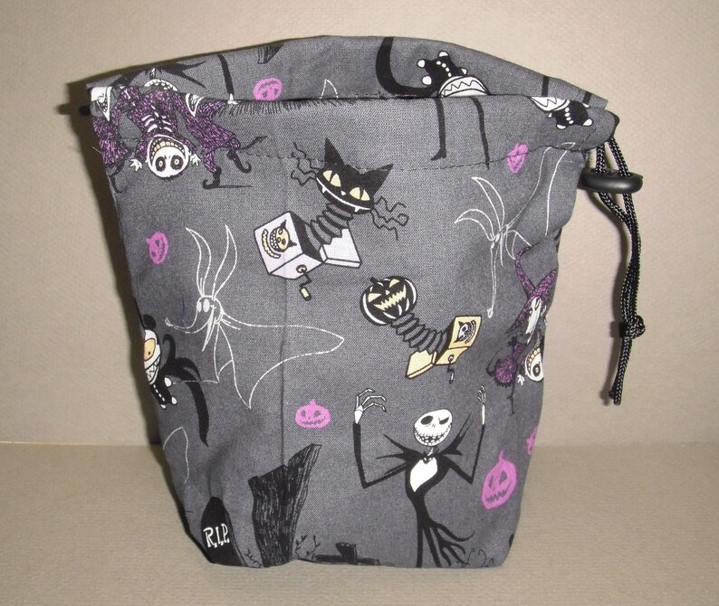 Puede incluir: Una bolsa de cord&oacute;n gris con un estampado en blanco y negro de personajes de Pesadilla antes de Navidad. La bolsa tiene un cord&oacute;n negro y un estampado morado y blanco de calabazas y murci&eacute;lagos.