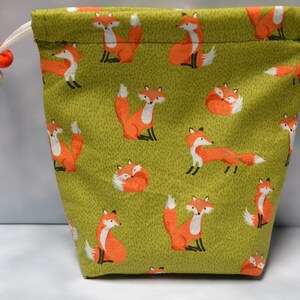 Peut inclure: Un sac à cordon vert avec un imprimé de renard. Le sac a un cordon blanc et un bouton rouge.
