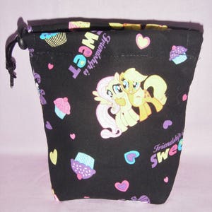 May include: Black drawstring bag with a My Little Pony theme. The bag features pink and yellow ponies, cupcakes, hearts, and the text "Friendship is Sweet".