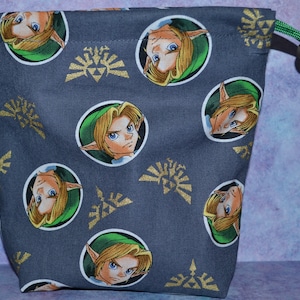 May include: A gray drawstring bag with a repeating pattern of a cartoon character with blonde hair and green clothing. The character is surrounded by a gold circle with a stylized design. The bag has a green drawstring with a black toggle.