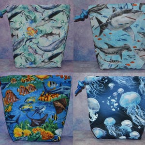 May include: Four drawstring bags with marine life patterns. One bag features whales and an octopus on a light blue background. Another displays sharks and fish on a blue backdrop. A third bag showcases colorful fish and coral. The last bag has jellyfish on a dark blue background.