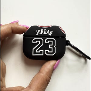 May include: Black AirPod case featuring the name "JORDAN" and the number "23" in white. The case has a red and white trim and a black carabiner clip.