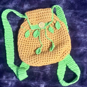 May include: A small brown crocheted backpack with green straps and leaf accents. The backpack has a single button closure.