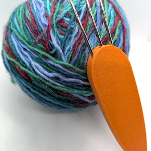 May include: A round ball of variegated yarn in shades of blue, green, and maroon, featuring a bright orange, carrot-shaped tool with three metal prongs inserted into the center of the yarn ball for knitting or crafting purposes.