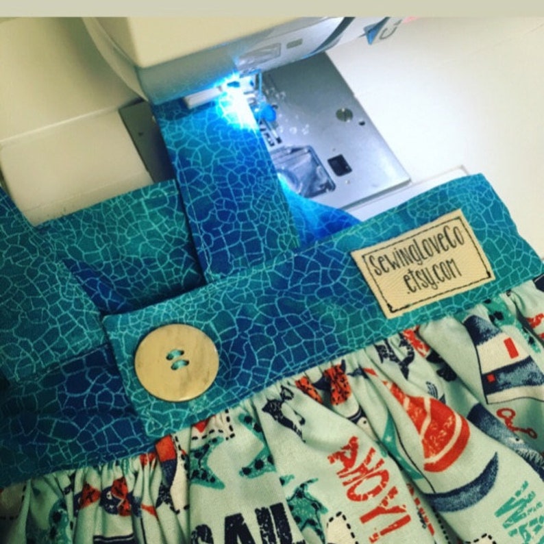 May include: A sewing machine is stitching a blue and green fabric with a button and a label that says "sewingloveco etsy.com".