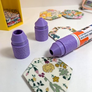 May include: Two purple 3D-printed glue stick caps with textured sides, a glue stick with a matching purple cap, and several fabric-covered hexagonal templates featuring floral patterns in yellow, pink, green, and blue, with a yellow storage bin.