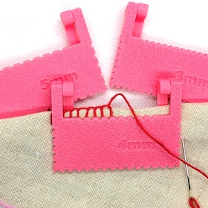 May include: Three bright pink plastic sewing tools with serrated edges, labeled 3mm, 4mm, and 5mm. One tool is positioned over a piece of beige fabric with red thread looped through the teeth, accompanied by a silver sewing needle.