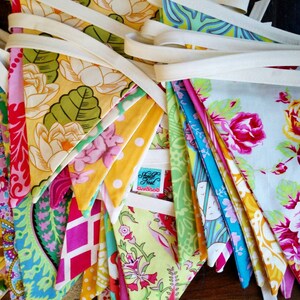 May include: A colorful fabric bunting with various floral and geometric patterns. The pennants are in shades of yellow, pink, green, and blue, connected by a cream-colored ribbon. The label reads "Scarlet Nest".