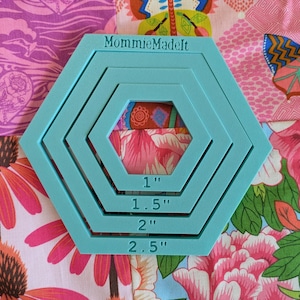 May include: A light blue plastic quilting template featuring four nested hexagonal frames. The top edge is embossed with the text MommieMadeIt. Each frame is clearly labeled with its respective size in inches: 1 inch, 1.5 inches, 2 inches, and 2.5 inches.