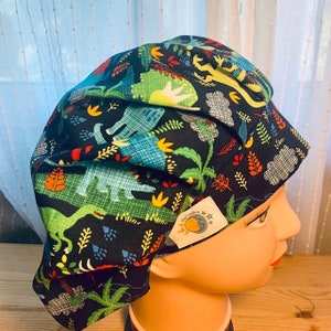 May include: A black scrub cap featuring a colorful dinosaur and plant pattern. The design includes green, blue, yellow, and red elements. A small white tag with text and a logo is sewn on the side. The cap is displayed on a mannequin head.