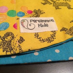 May include: Yellow fabric with a black and white design of people and animals. The fabric has a white label with the text "Persimmon Kids" in black lettering.