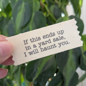 May include: A rectangular off-white fabric label with jagged, pinked edges featuring the black printed text: If this ends up in a yard sale, I will haunt you. The label has a visible woven texture and is held against a background of green leaves.